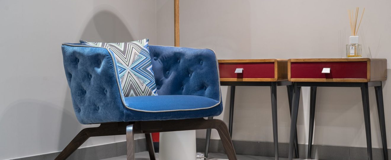 Comfortable blue armchair with cushion near lamp and wooden tables with diffusor in white room